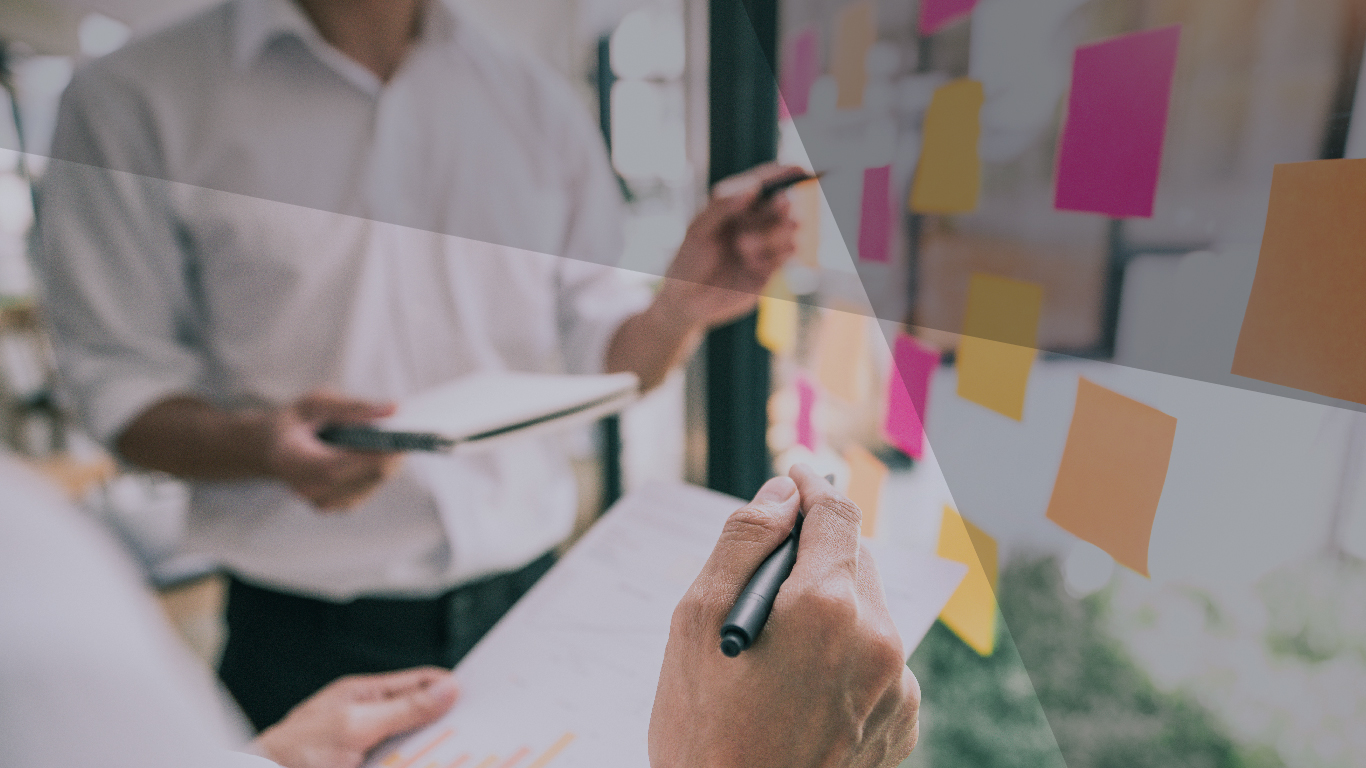 Two people with a pen and pad working together on a board with sticky notes scattered across the board